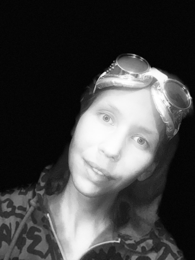 Black and white portrait of a woman in partial smile wearing silver goggles on her forehead and a patterned hoodie.