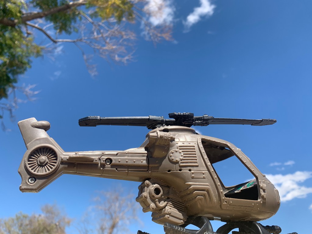 Toy plastic helicopter posed against the clear blue sky punctuated by a few trees and a spatter of whisky clouds
