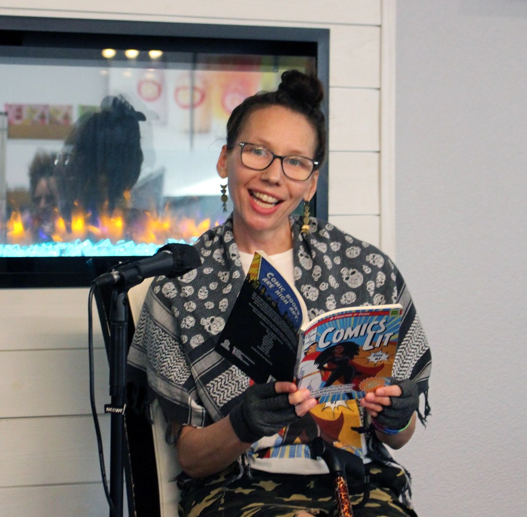 Ada sits behind a mic with a paperback copy of comics lit. SHe is wearing a black and white skull keffia, white tee, black gloves, and camo skirt. She is looking at the camera, smiling, mid-sentence reading from the book. There is a leopard print cane balanced in front of her. 